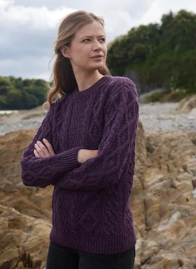 woman standing in rocky beach wearing purple aran sweater with arms folded