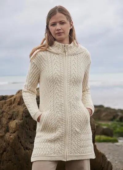 brown haired woman standing on rocky beach wearing a cream aran coatigan with hands in pockets