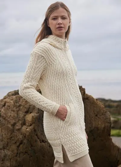 side angle of brown haired woman standing on rocky beach wearing a cream aran coatigan with hands in pockets