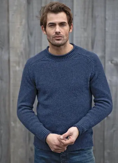man standing against grey fence wearing blue knit sweater with clasped hands