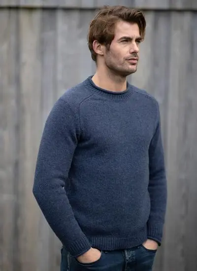 man standing against grey fence wearing blue knit sweater with hands in pockets