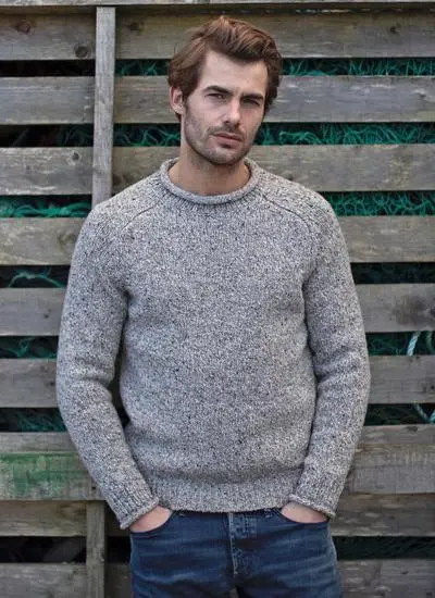 man standing against grey wooden fence wearing grey sweater with hands in pockets