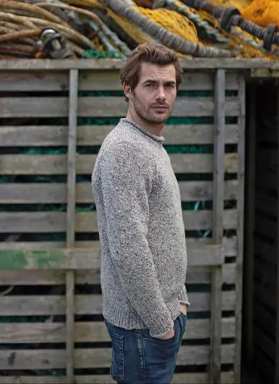 side angle shot of man standing against grey wooden fence wearing grey sweater with hands in pockets