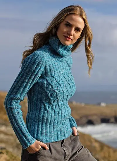 blonde woman standing by the sea on sunny day wearing blue sweater with hand in pocket