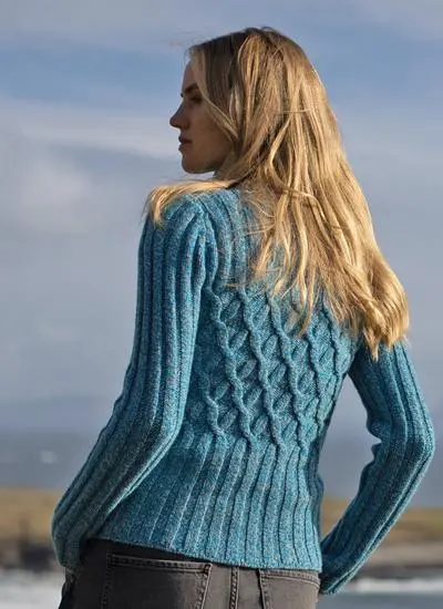 back shot of blonde woman standing by the sea on sunny day wearing blue sweater with hand in pocket