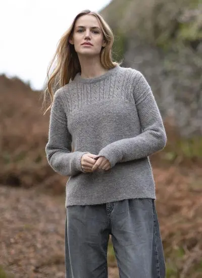 blonde woman standing in rocky outdoor area wearing a grey eyelet knit sweater with hands clasped