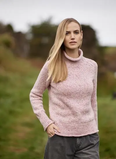 blonde woman standing in grassy outdoor area wearing light pink sweater with one hand in pocket