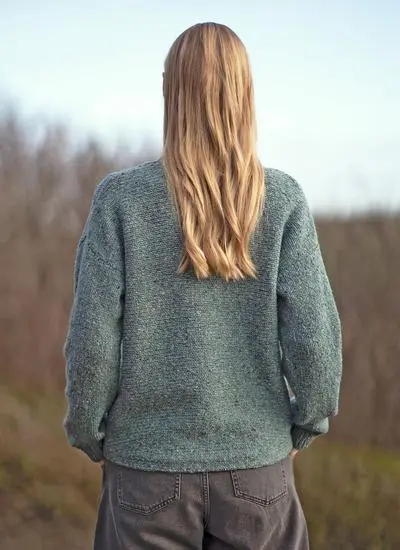 back shot of a woman with blonde hair standing outside with hands in pockets wearing jade fisherman knitted sweater