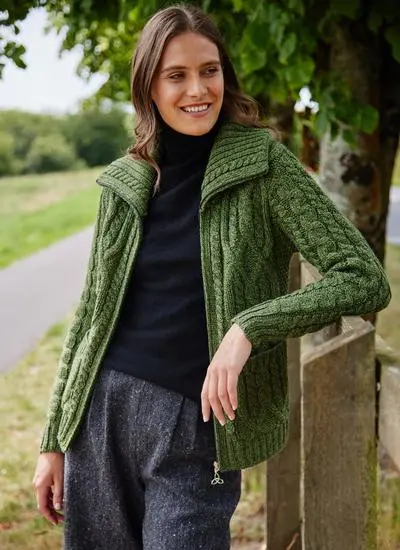 smiling brown haired woman leaning against wooden fence in park wearing an open green zip up aran cardigan with black top underneath