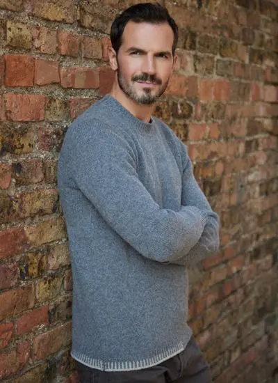 brunette man wearing grey sweater standing by red brick wall with arms folded