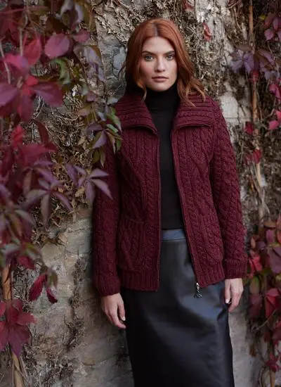 red haired woman standing against wall with red vines wearing deep red aran zip cardigan 