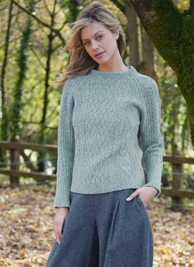 A woman in a green Aran sweater and gray trousers stands in a wooded area, with autumn leaves in the background.