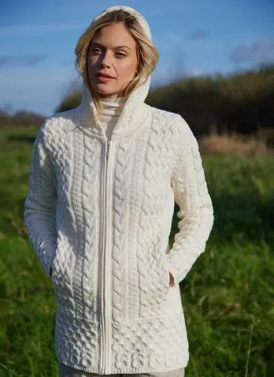 A blonde woman standing in field wearing celtic aran natural hoody, hood up, hands in pockets