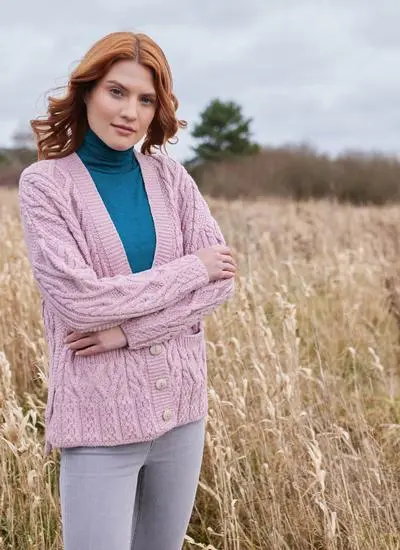 Red hair lady wearing a pink aran cardigan, with a blue polo neck under it, in a field with her arms folded
