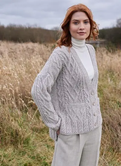 red haired woman standing in field of reeds wearing a grey aran cardigan with white polo beck underneath and hands in pockets