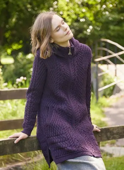 woman with short brown hair in wooded area leaning against wooden fence wearing purple aran coatigan