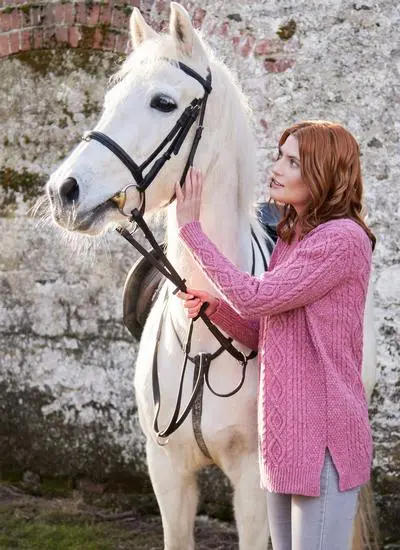 red haired woman wearing pink turtle neck sweater in stables petting white horse